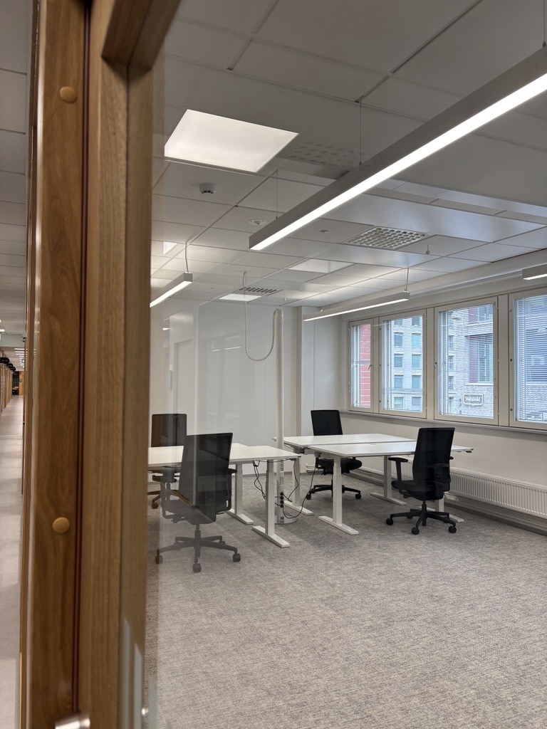 Näkymä moderniin toimistoon, jossa on suuret ikkunat, valkoiset työpöydät ja mustat pyörivät työtuolit. Huone on valaistu kattovalaisimilla, ja sen lattia on peitetty harmaalla matolla. Käytävä ja puinen ovikarmit näkyvät osittain.