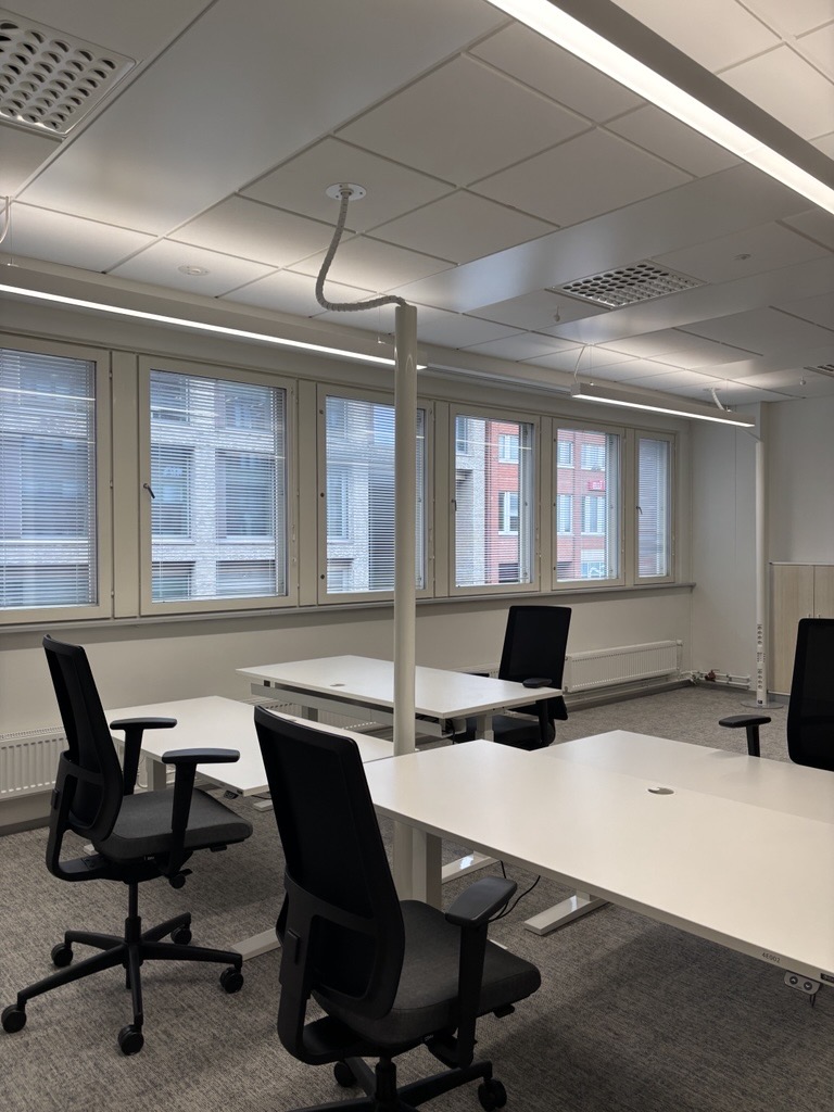 Moderni toimisto, jossa on useita tyhjiä työpöytiä ja mustia tuoleja, suuret ikkunat, joissa on kaihtimet, harmaa matto ja kattovalaisimet. Huone on valoisa, ja ikkunoista avautuu näkymä muihin rakennuksiin.