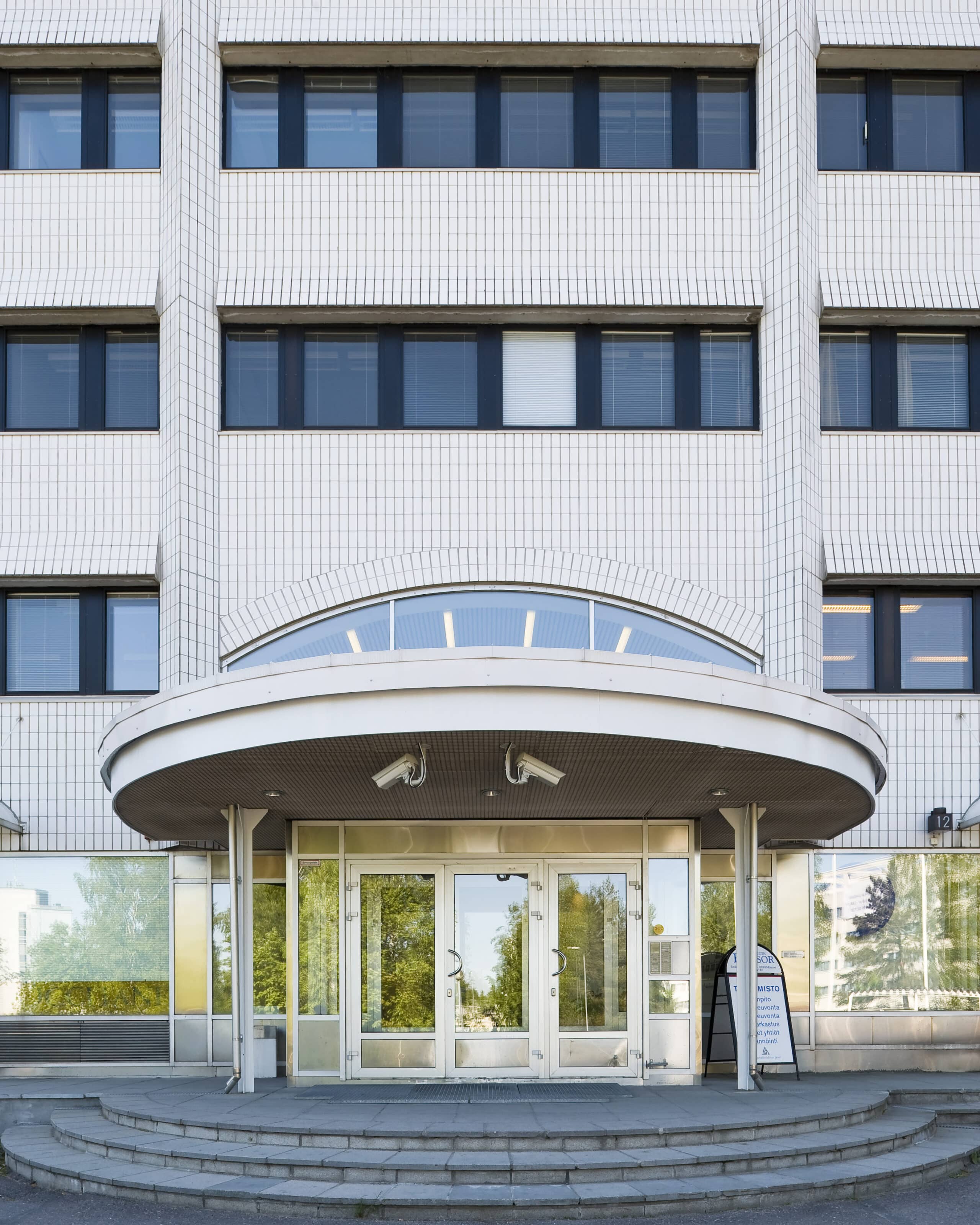 Kuvassa näkyy modernin valkoisen rakennuksen pääsisäänkäynti, jossa on suuret ikkunat, lasiovet, kaareva katos ja sisäänkäyntiin johtavat portaat. Puut heijastuvat lasista.