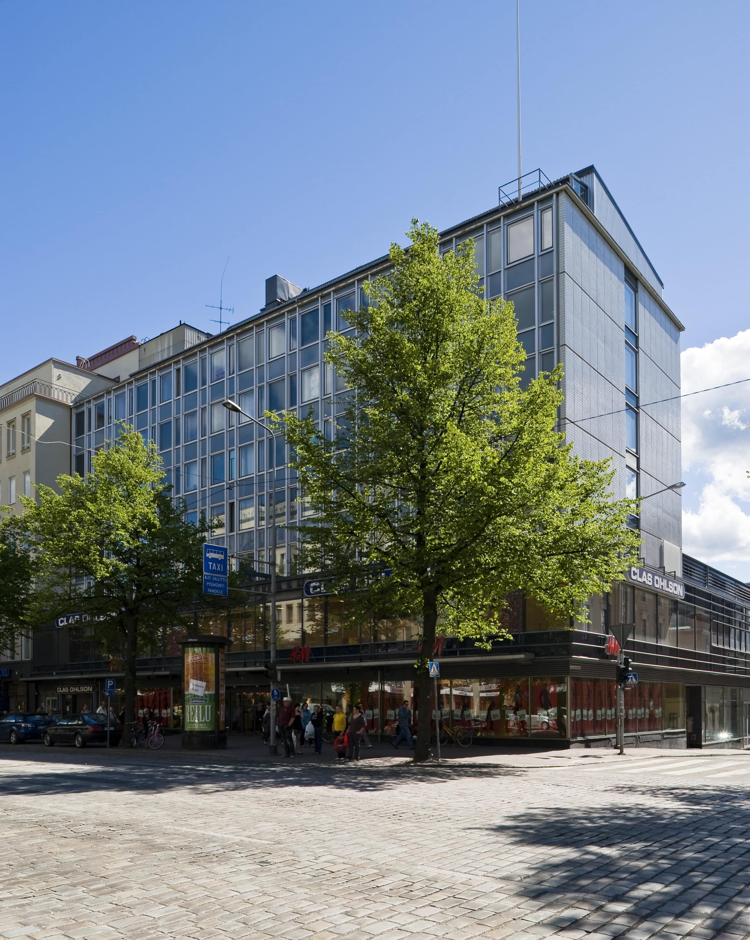 Moderni lasinen toimistorakennus, jonka edessä on puita, sijaitsee mukulakivikadun kulmassa kirkkaan sinisen taivaan alla. Jalankulkijat kulkevat jalkakäytävällä.