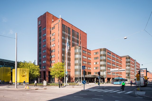 Vilkkaalla kaupunkikadulla seisoo korkea, punatiilinen rakennus, jossa on paljon ikkunoita. Ihmiset kävelevät ja pyöräilevät sen läheisyydessä, ja risteyksessä pysähtyy sininen bussi. Taivas on kirkas ja sininen.