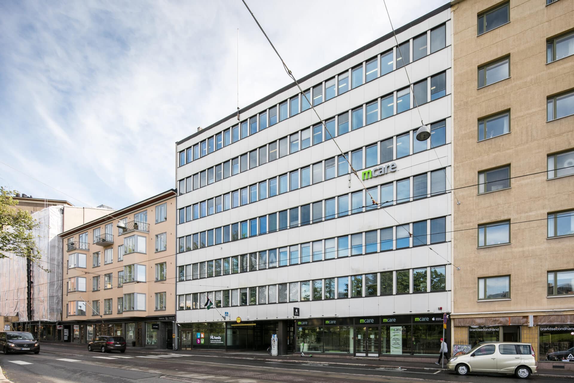 Näkymä kadulle, jossa näkyy moderneja ja vanhempia kerrostaloja sekä valkoinen keskikorkea toimistorakennus, jossa on Mate-logo, aurinkoisena päivänä sinisen taivaan alla.