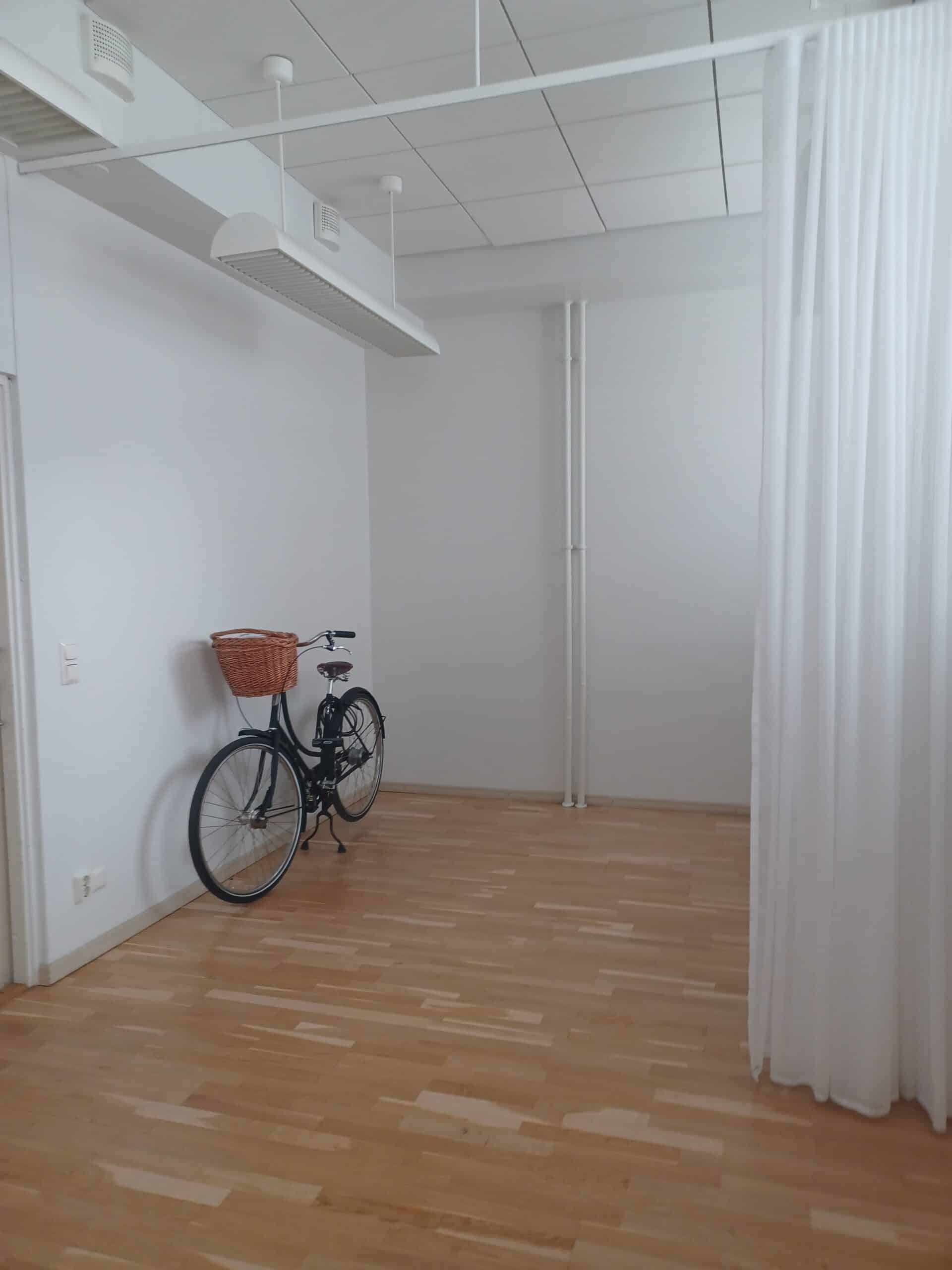 Musta polkupyörä, jossa on korikori, on pysäköity valkoista seinää vasten minimalistisessa, tyhjässä huoneessa, jossa on vaaleat puulattiat ja valkoiset verhot oikealla puolella.