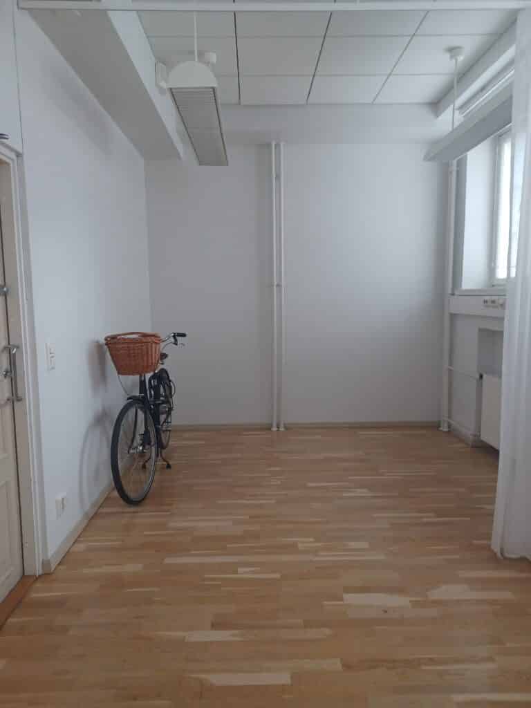 Pääosin tyhjä, valoisa huone, jossa on puulattia ja valkoiset seinät, seinää vasten seisoo polkupyörä, jossa on korikori.