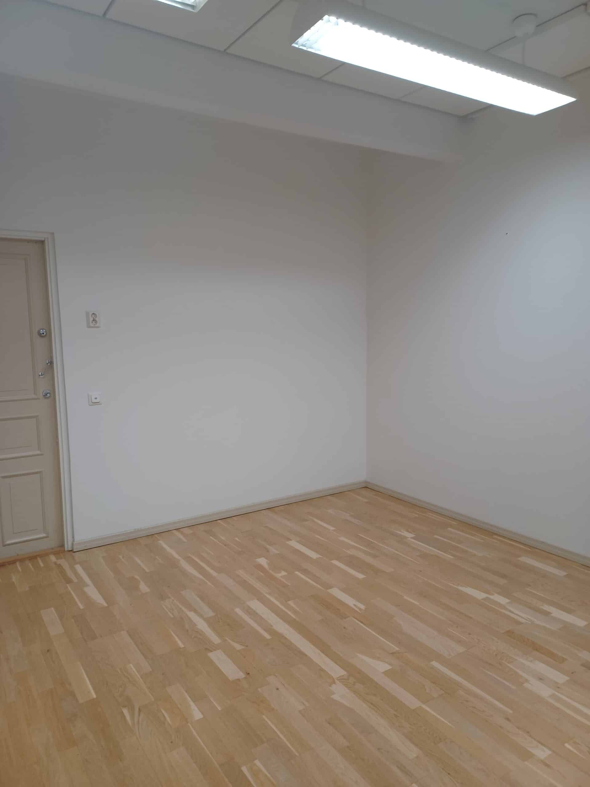 Tyhjä huone, jossa on vaaleat puulattiat, valkoiset seinät, suljettu beige ovi ja loisteputkivalaisimet katossa.
