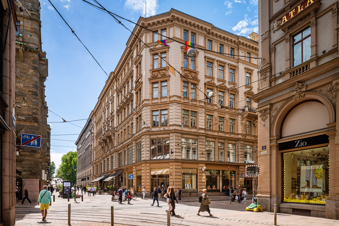 Ihmiset kävelevät aurinkoisella kaupunkikadulla, jota reunustavat historialliset rakennukset, kaupat ja kahvilat. Kulmarakennuksesta roikkuu lippuja, ja kadun keskellä kulkee raitiotiekiskoja.