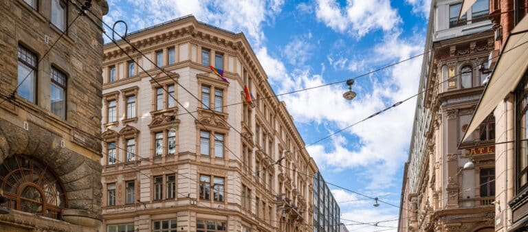 Historialliset kivirakennukset, joissa on paljon ikkunoita, reunustavat kaupungin katua osittain pilvisen sinisen taivaan alla. Yläpuolella roikkuvat sähköjohdot ja katuvalot, ja kulmarakennuksessa näkyy kaksi sateenkaarilippua.