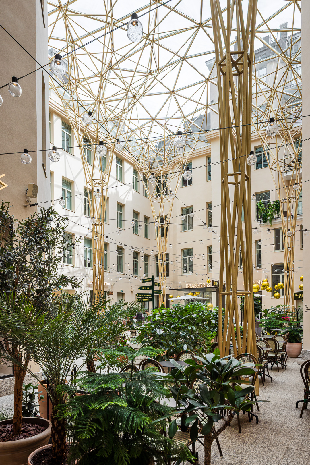 Valoisa atrium, jossa on ruukkukasveja, kahvilapöytiä ja tuoleja, ja jota ympäröivät modernit kermanväriset rakennukset. Katossa on geometrinen kultainen metallirakenne, josta roikkuu valaisimia.