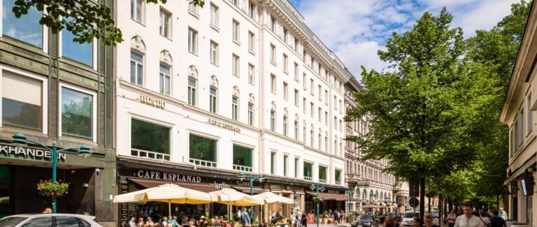 Katuvaikutelma, jossa ihmiset ruokailevat Café Esplanadin ulkoterassilla; kahvila sijaitsee valkoisen monikerroksisen rakennuksen pohjakerroksessa, ja taustalla näkyy puita ja sininen taivas.