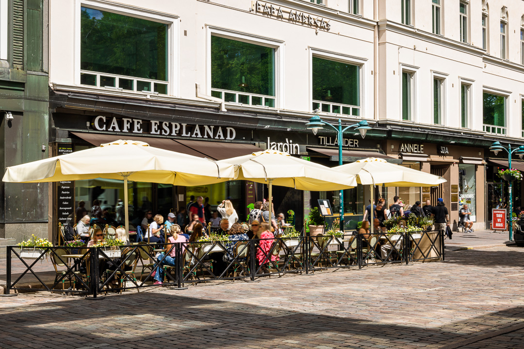 Aurinkoisena päivänä ihmiset istuvat pöydissä sateenvarjojen alla Café Esplanadin ulkoterassilla. Kahvila sijaitsee vaaleanvärisen, suurilla ikkunoilla varustetun rakennuksen pohjakerroksessa, ja ulkoterassin reunaa kiertävät kukat.