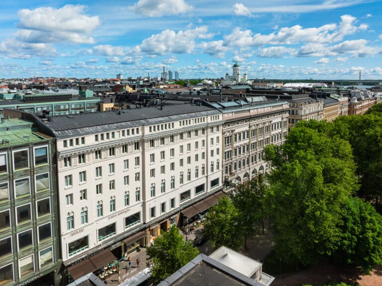Ilmakuva kaupungista, jossa on perinteisiä valkoisia rakennuksia, etualalla vihreitä puita ja yläpuolella sininen taivas, jossa on hajanaisia pilviä. Etäisyydessä kohoaa valkoisella kupolilla varustettu katedraali.