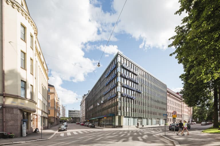 Moderni lasinen toimistorakennus kaupungin kadunkulmassa, jota ympäröivät vanhemmat rakennukset ja puut; kadunvarsilla on pysäköityjä autoja, ja muutamat ihmiset kävelevät jalkakäytävillä osittain pilvisen taivaan alla.