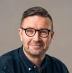 Mies, jolla on lyhyet, siististi muotoillut hiukset ja joka käyttää pyöreitä mustia silmälaseja sekä tummaa napitettua paitaa, hymyilee lempeästi kameraan. Tausta on yksinkertainen ja neutraali.
