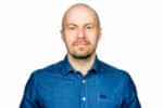 Mies, jolla on ajeltu pää, lyhyt parta ja viikset, on pukeutunut siniseen napitukseen ja katsoo kameraan ilmeettömästi yksinkertaista valkoista taustaa vasten.