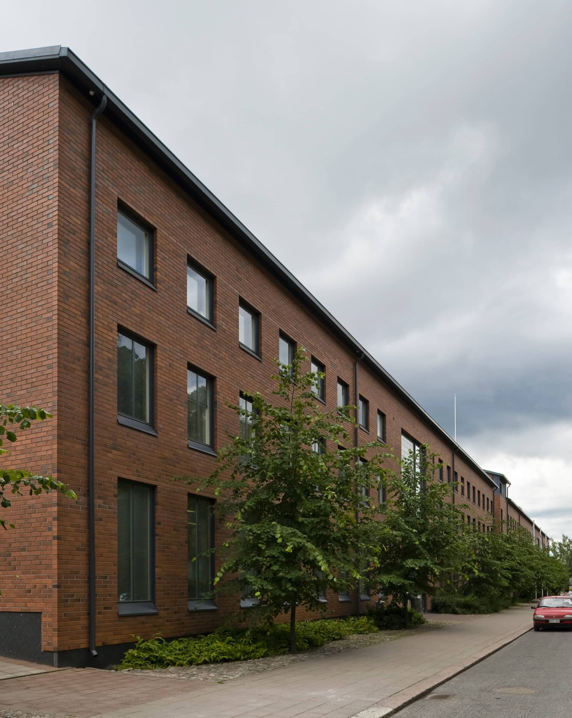 Hiljaisen kadun varrella seisoo moderni, punatiilinen rakennus, jossa on suuret ikkunat. Pilvisen taivaan alla jalkakäytävän varrella kasvaa pieniä puita ja pensaita.