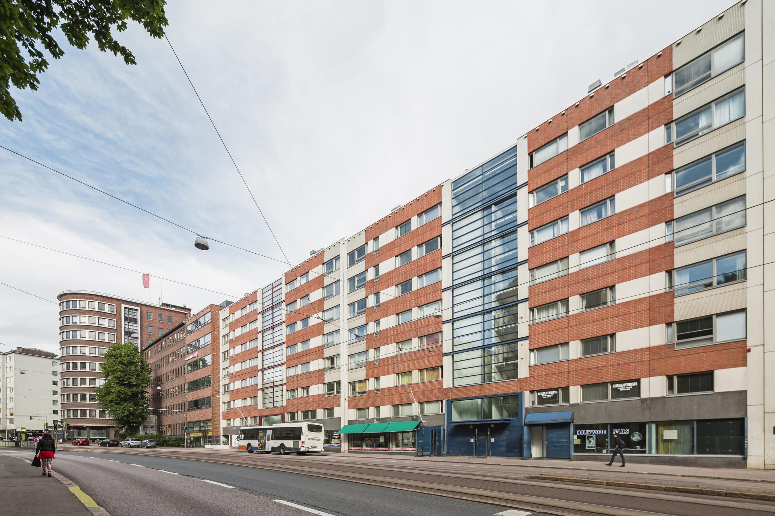 Kaupunkikatu, jolla on moderneja, monikerroksisia kerrostaloja, joiden julkisivut ovat punavalkoisia, lasi-ikkunat, katutasossa sijaitsevia kauppoja ja ohi ajava bussi. Jalankulkijat kulkevat jalkakäytävällä osittain pilvisen taivaan alla.
