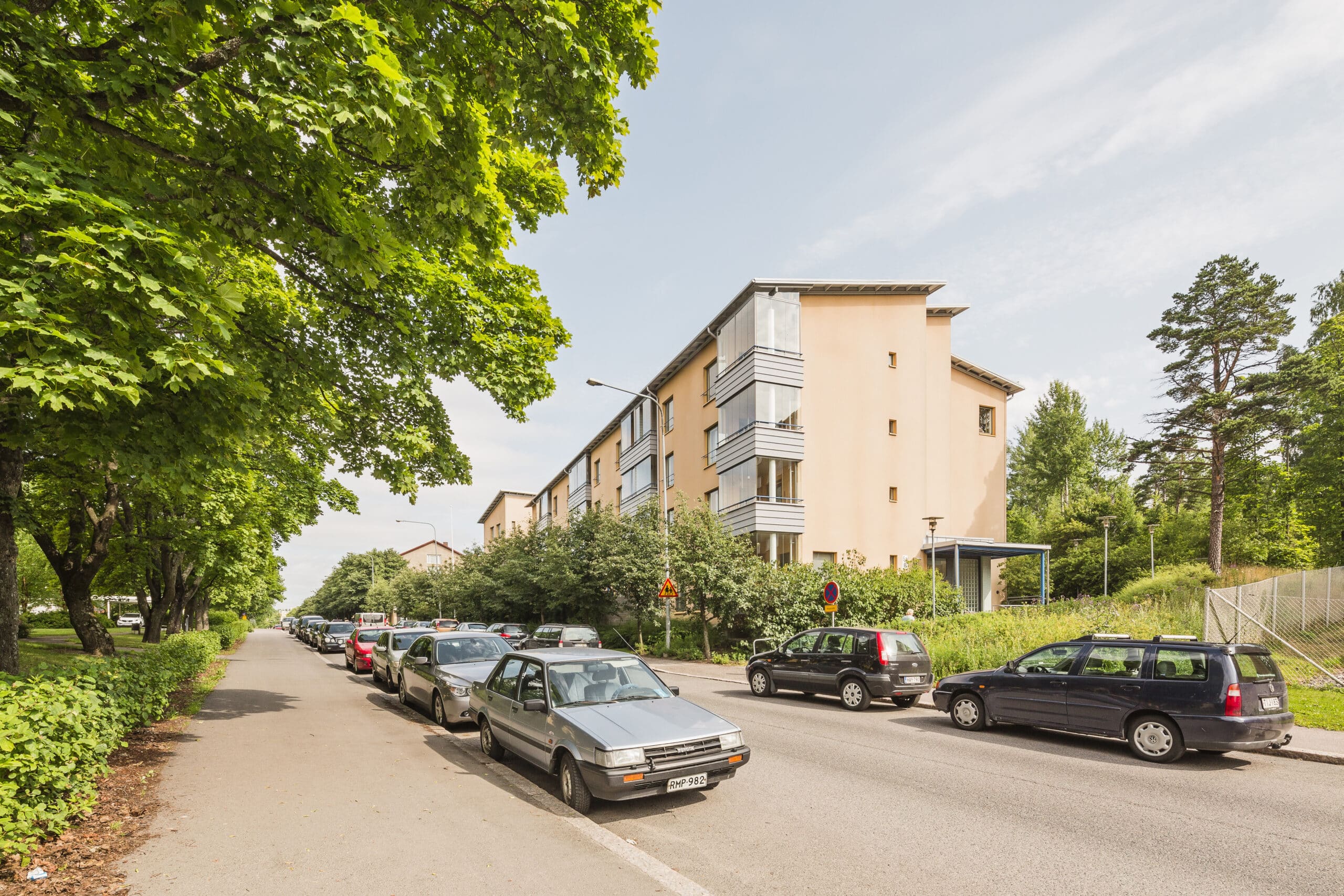 Pysäköityjen autojen rivi reunustaa katua vaalean persikanväristen seinien omaavan kerrostalon vieressä; rakennusta ympäröivät vihreät puut ja pensaat osittain pilvisen taivaan alla.