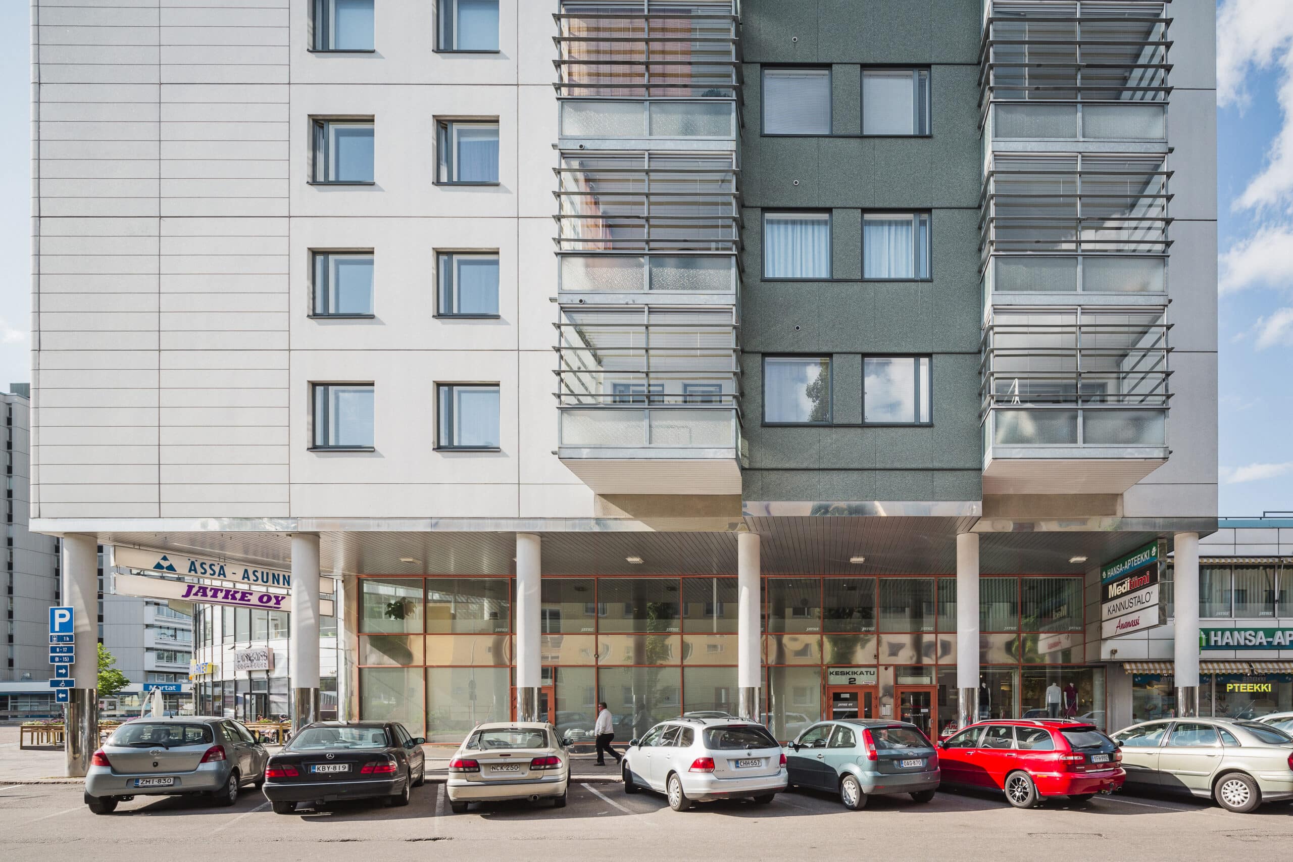 Moderni rakennus, jossa on suuret ikkunat ja lasinen sisäänkäynti, useita liiketiloja katutasossa sekä kuusi pysäköityä autoa edessä. Taivas on kirkas ja sininen.