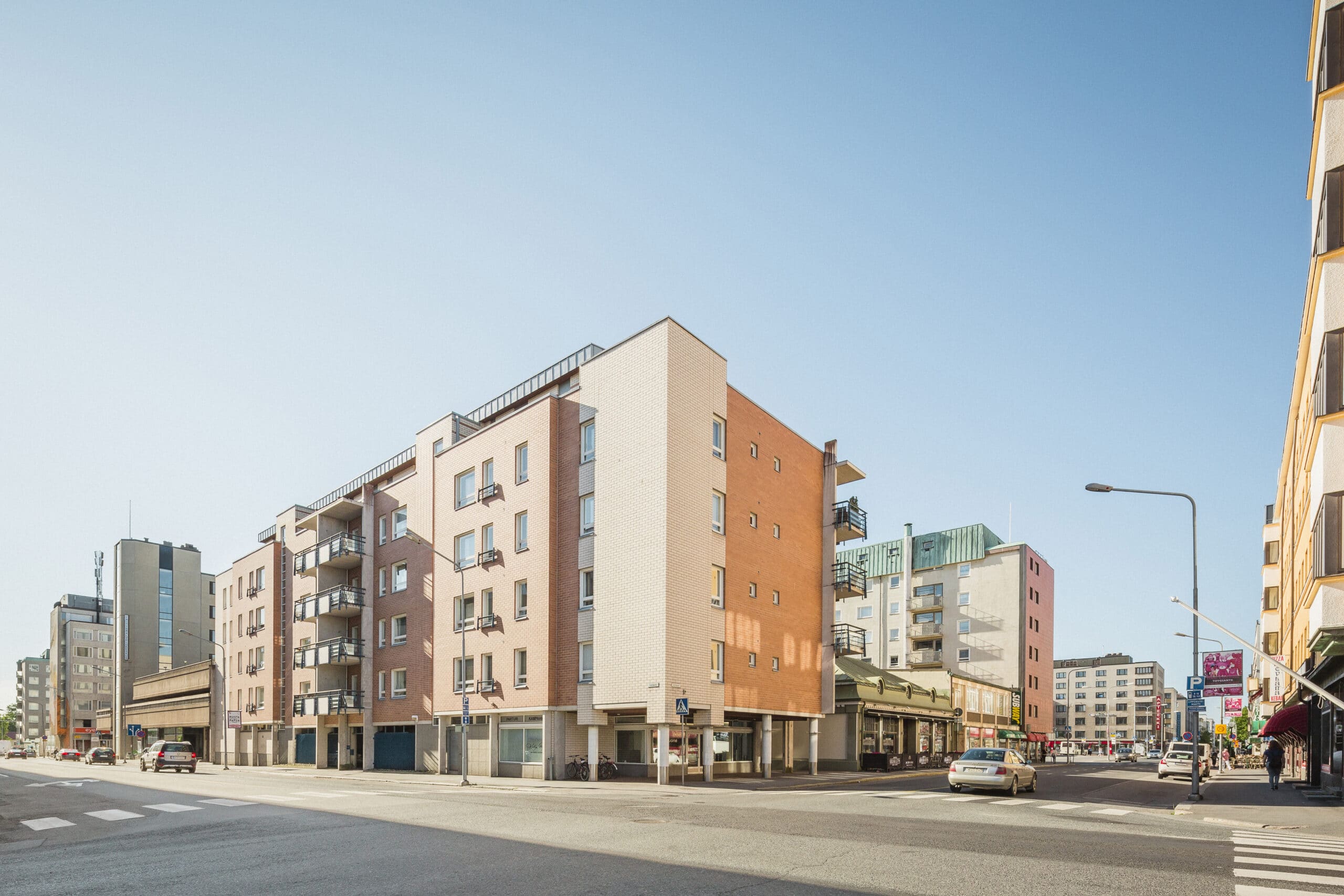 Kaupungin katu, jolla on moderneja monikerroksisia kerrostaloja, katutasossa sijaitsevia kauppoja, kirkas sininen taivas sekä taustalla muutama jalankulkija ja auto.