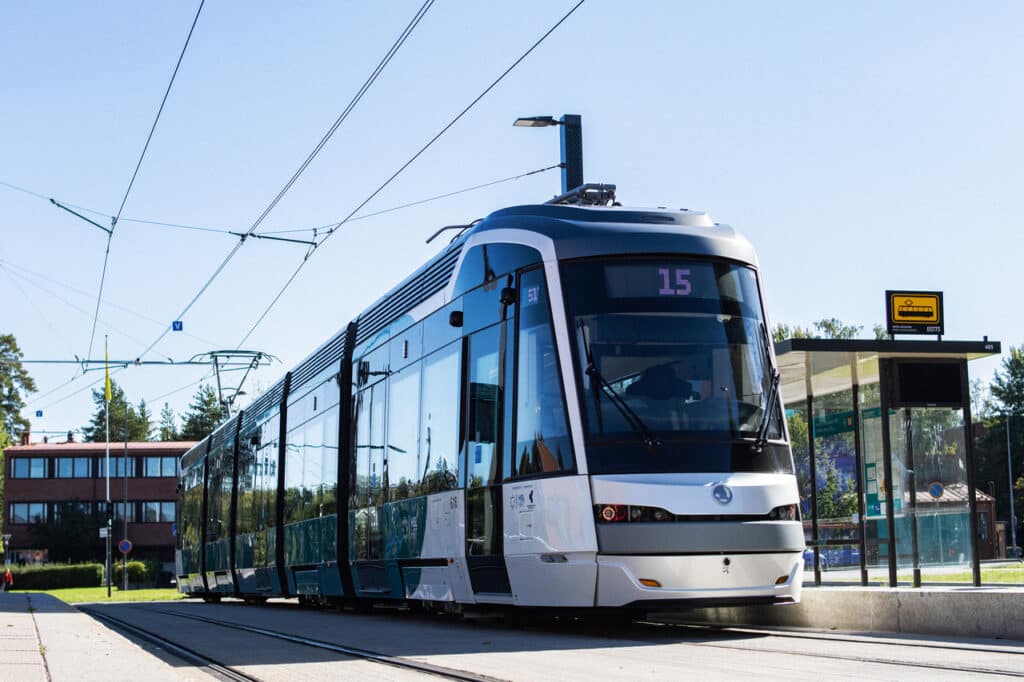 Moderni raitiovaunu numero 15 pysähtyy aurinkoisella ulkoasemalla, jossa on lasikatokset. Taustalla näkyy yläjohdot ja muutamia rakennuksia.