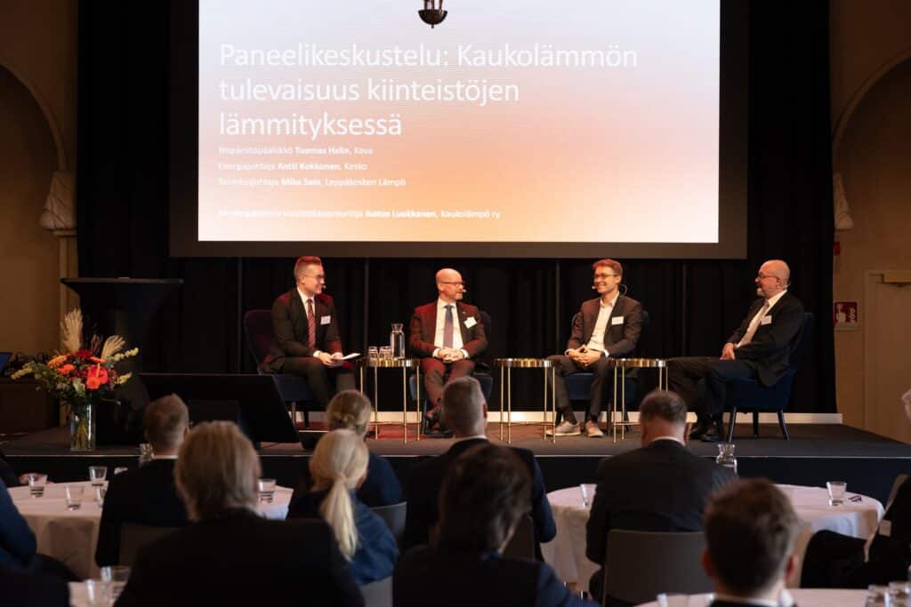 Neljä liikeasuisia miestä istuu lavalla paneelikeskustelussa, jonka otsikko on ”Paneelikeskustelu: Kaukolämmön tulevaisuus kiinteistöjen lämmityksessä”, konferenssisalissa olevan yleisön edessä.