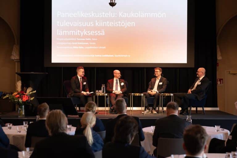 Neljä liikeasuisia miestä istuu lavalla paneelikeskustelussa, jonka otsikko on ”Paneelikeskustelu: Kaukolämmön tulevaisuus kiinteistöjen lämmityksessä”, konferenssisalissa olevan yleisön edessä.